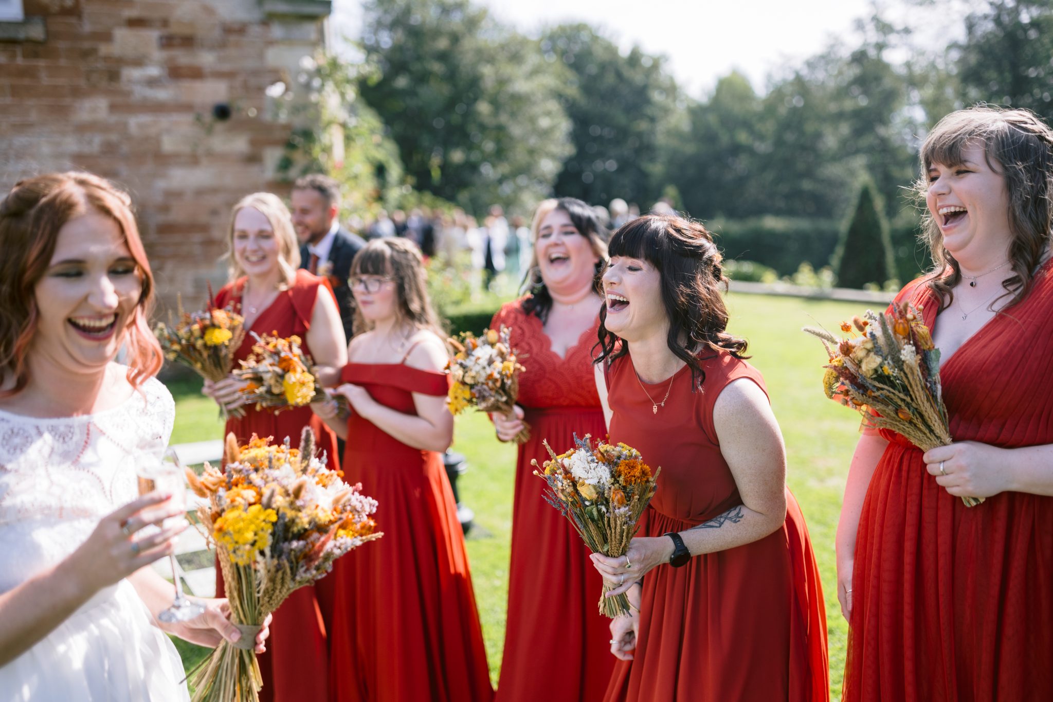 A group of women in red dresses, holding bouquets, are laughing together outdoors at a beautiful summer marquee wedding at Kirklinton Hall.