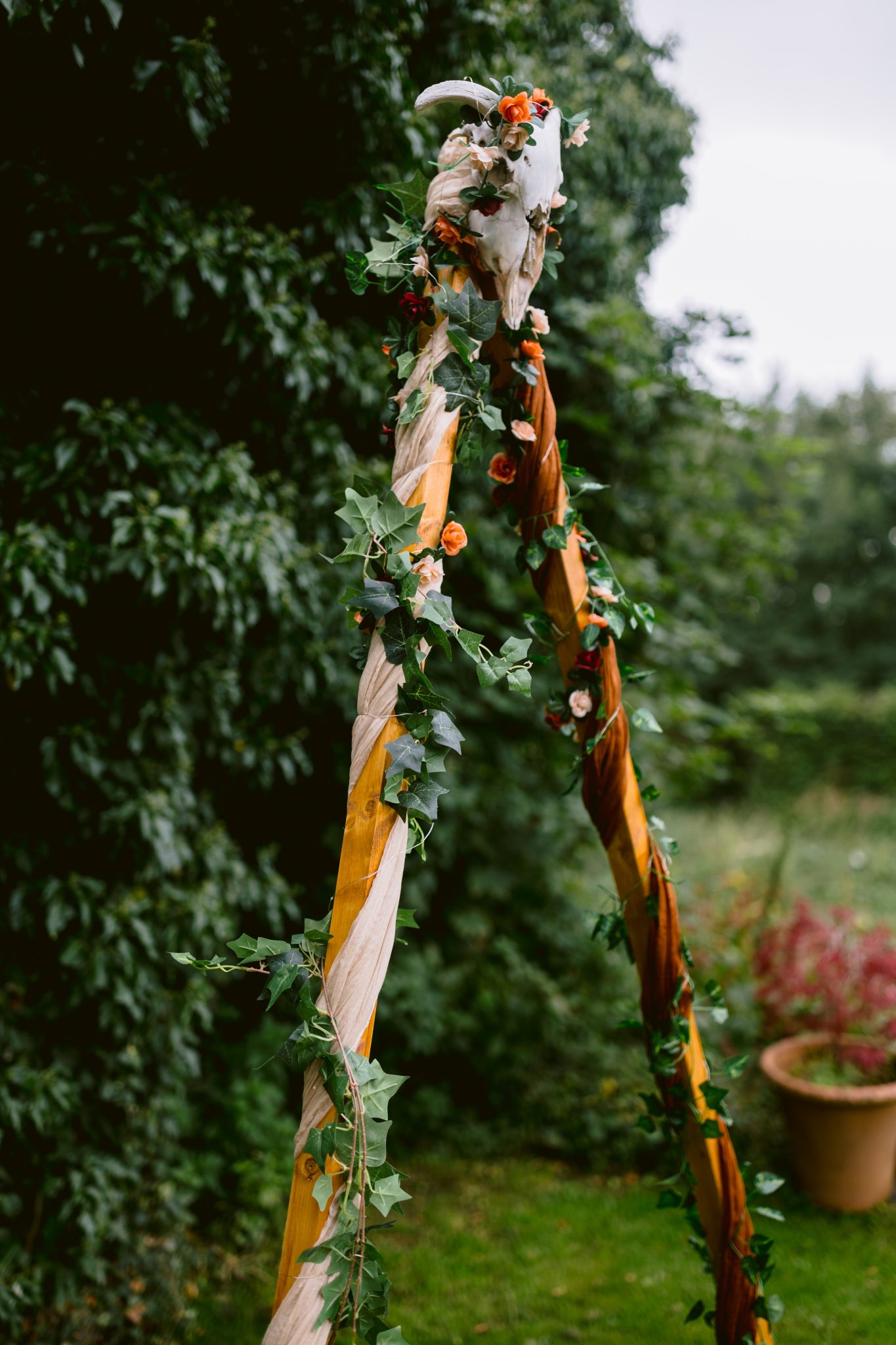 A stunning wooden arch at Kirklinton Hall, gracefully adorned with green ivy and vibrant orange flowers, crowned by a white skull. Set in enchanting gardens with summer trees as a backdrop, it exudes an air of natural elegance perfect for any marquee wedding.