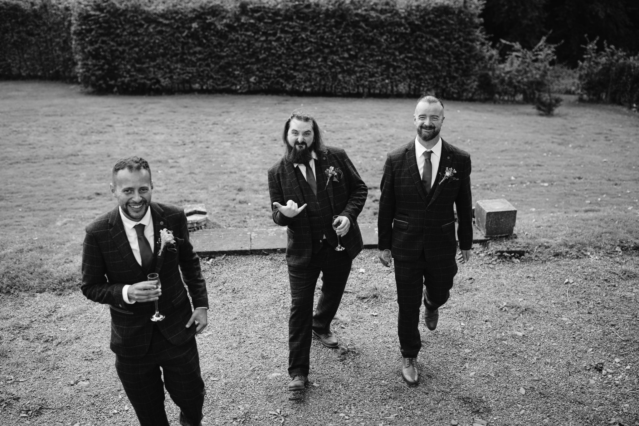 Three men in suits stroll along a gravel path, drinks in hand and smiles bright. Behind them, the lush gardens of Kirklinton Hall provide a perfect backdrop. This black and white image captures the timeless elegance often seen at a summer marquee wedding.