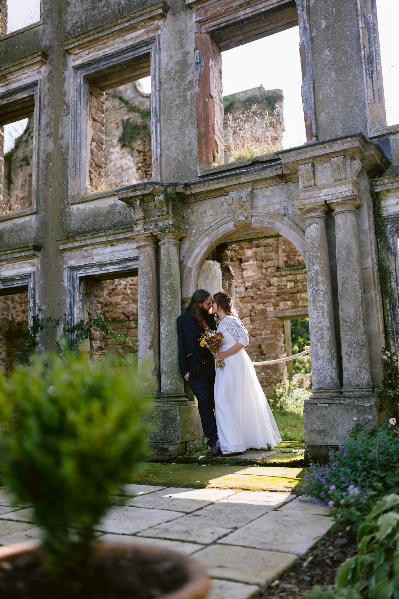 A couple in wedding attire stands intimately in a stone archway of an old, ruined building at Kirklinton Hall. A potted plant is in the foreground, hinting at the lush gardens that surround this enchanting venue perfect for a summer marquee wedding.