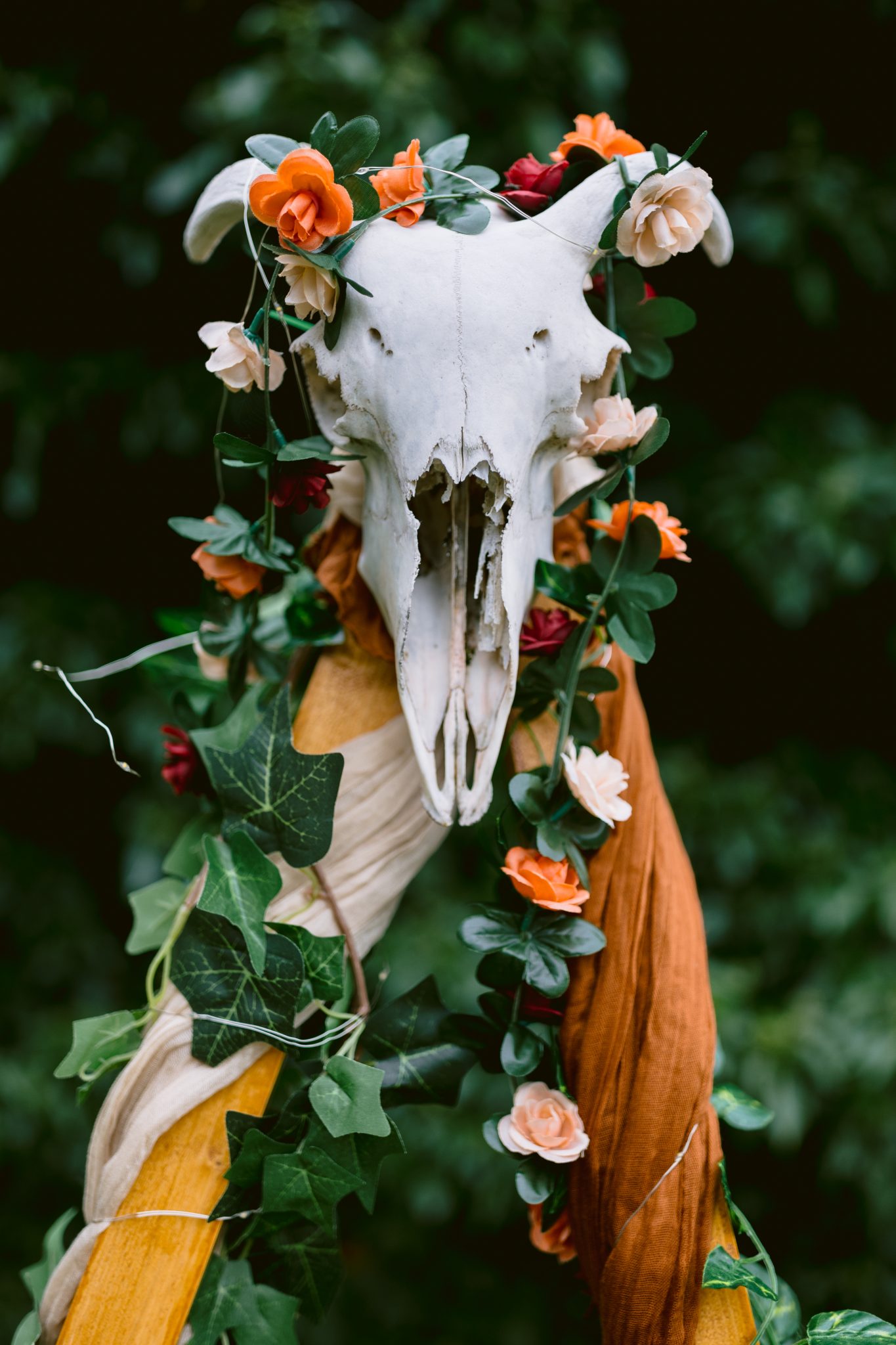 An animal skull adorned with orange and pink flowers, green leaves, and draped fabric sits elegantly amidst Kirklinton Hall gardens, set against a dark green foliage background.