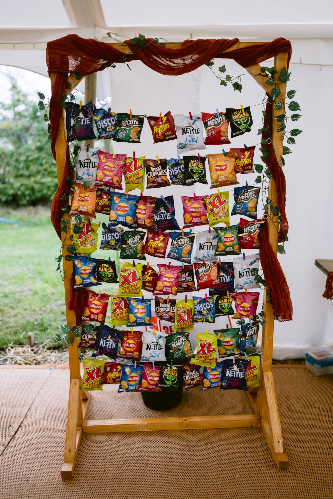 A wooden stand at Kirklinton Hall proudly displays a variety of snack packs, like crisps and chips, arranged in rows. The setup is elegantly adorned with red cloth and leafy vines, evoking the charm of a summer marquee wedding nestled in picturesque gardens.