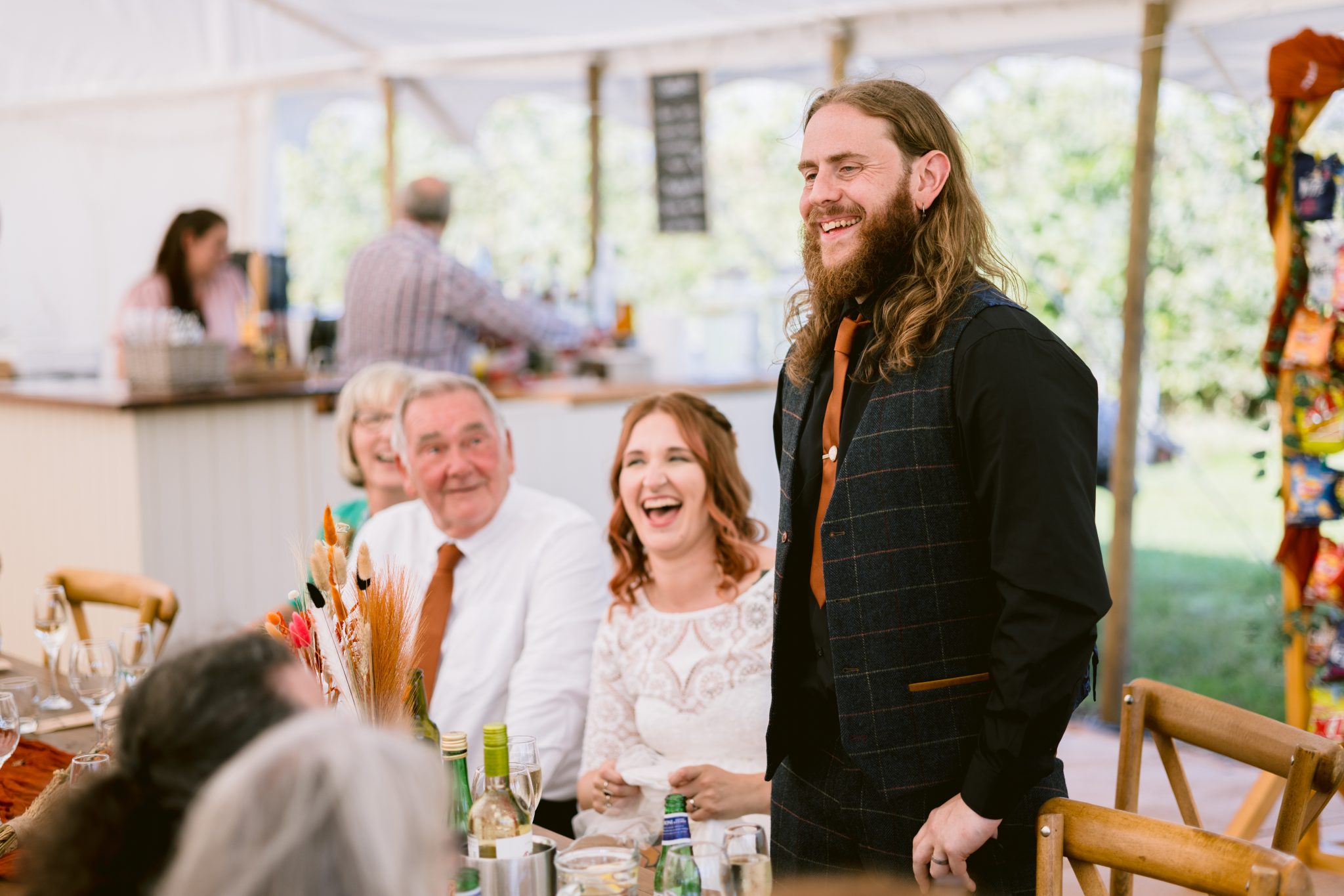 A man with long hair stands smiling and talking at a table during a wedding at Kirklinton Hall. Guests seated around him, including a laughing woman, are engaged and attentive, enjoying the charming Gardens of this outdoor gathering.