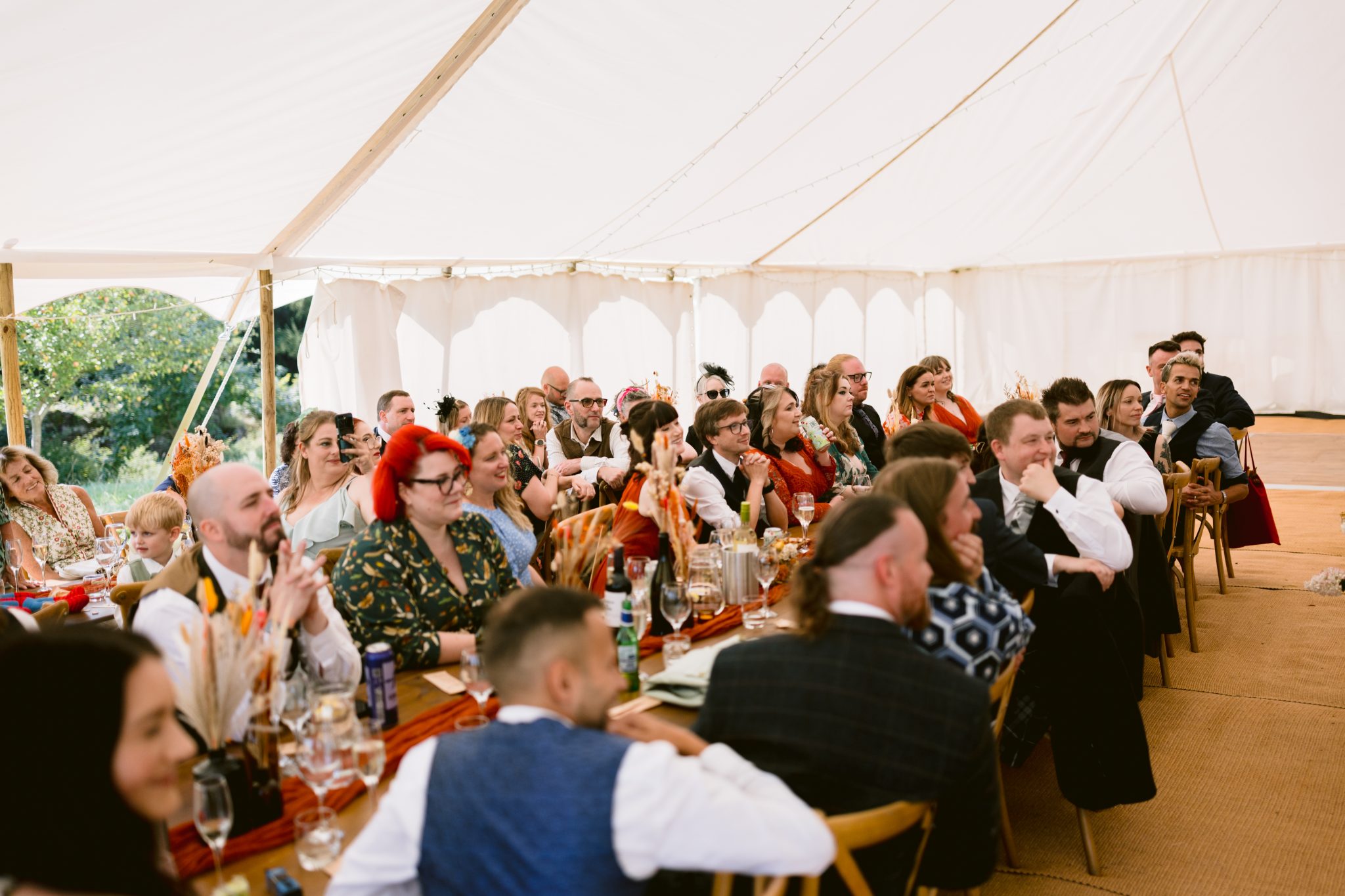Guests seated under a large white tent at a Summer Marquee Wedding, listening attentively. The picturesque setting of Kirklinton Hall's gardens enhances the ambiance, with tables elegantly set with drinks and decorations.