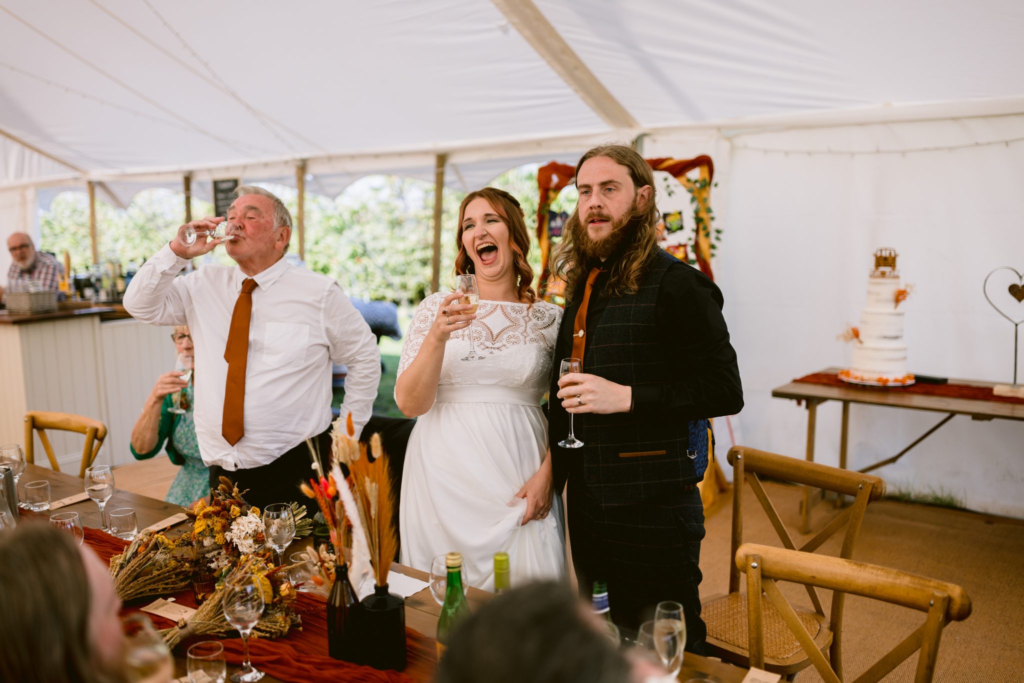 The bride and groom, elegantly dressed for their summer marquee wedding at Kirklinton Hall, smile while holding champagne glasses amidst the gardens with guests gathered around under the tent.