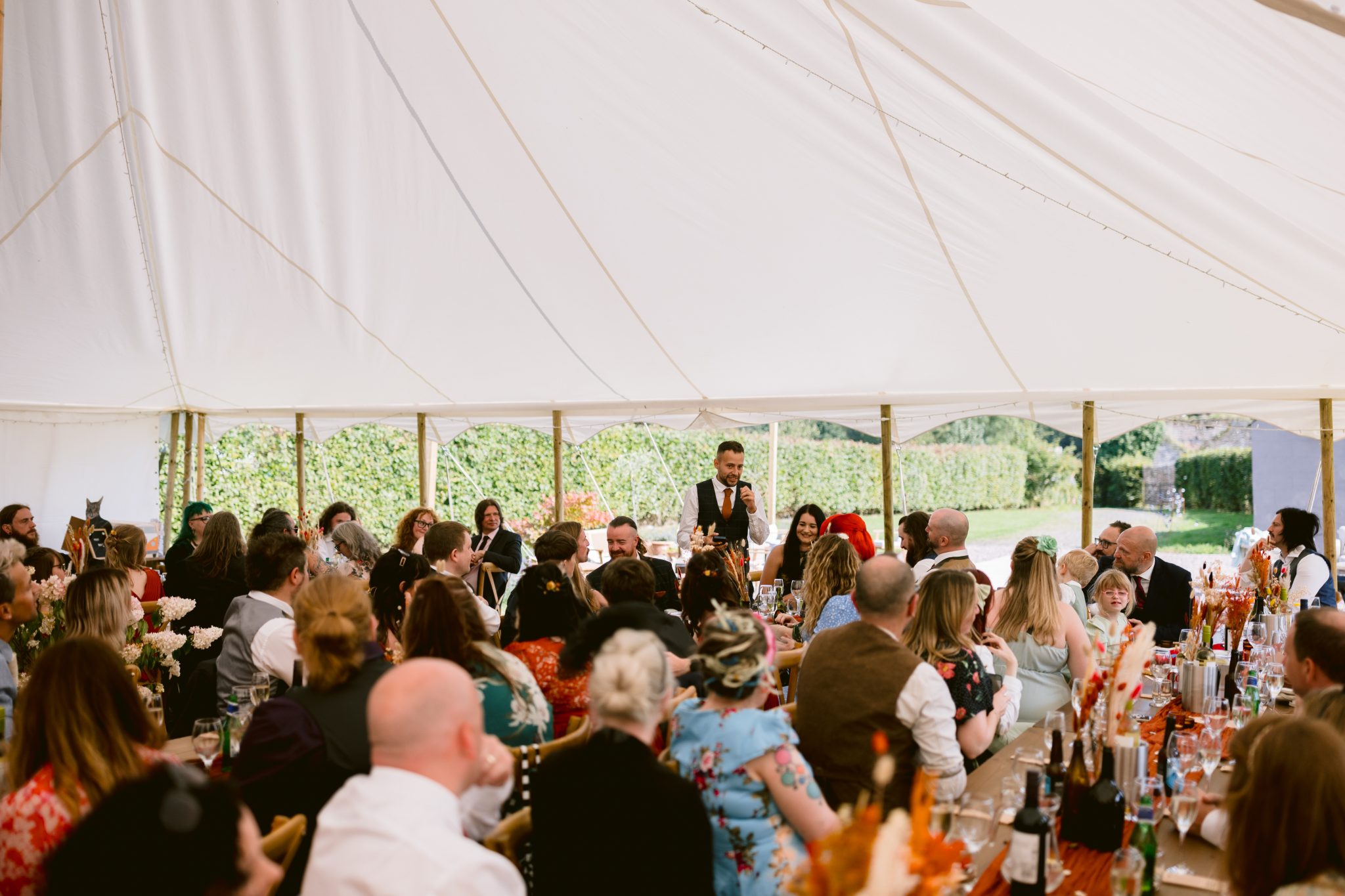 A person stands and speaks to a large group of seated guests inside a white tent, decorated with flowers and dining setups, at a beautiful Summer Marquee Wedding beside the lush gardens of Kirklinton Hall.