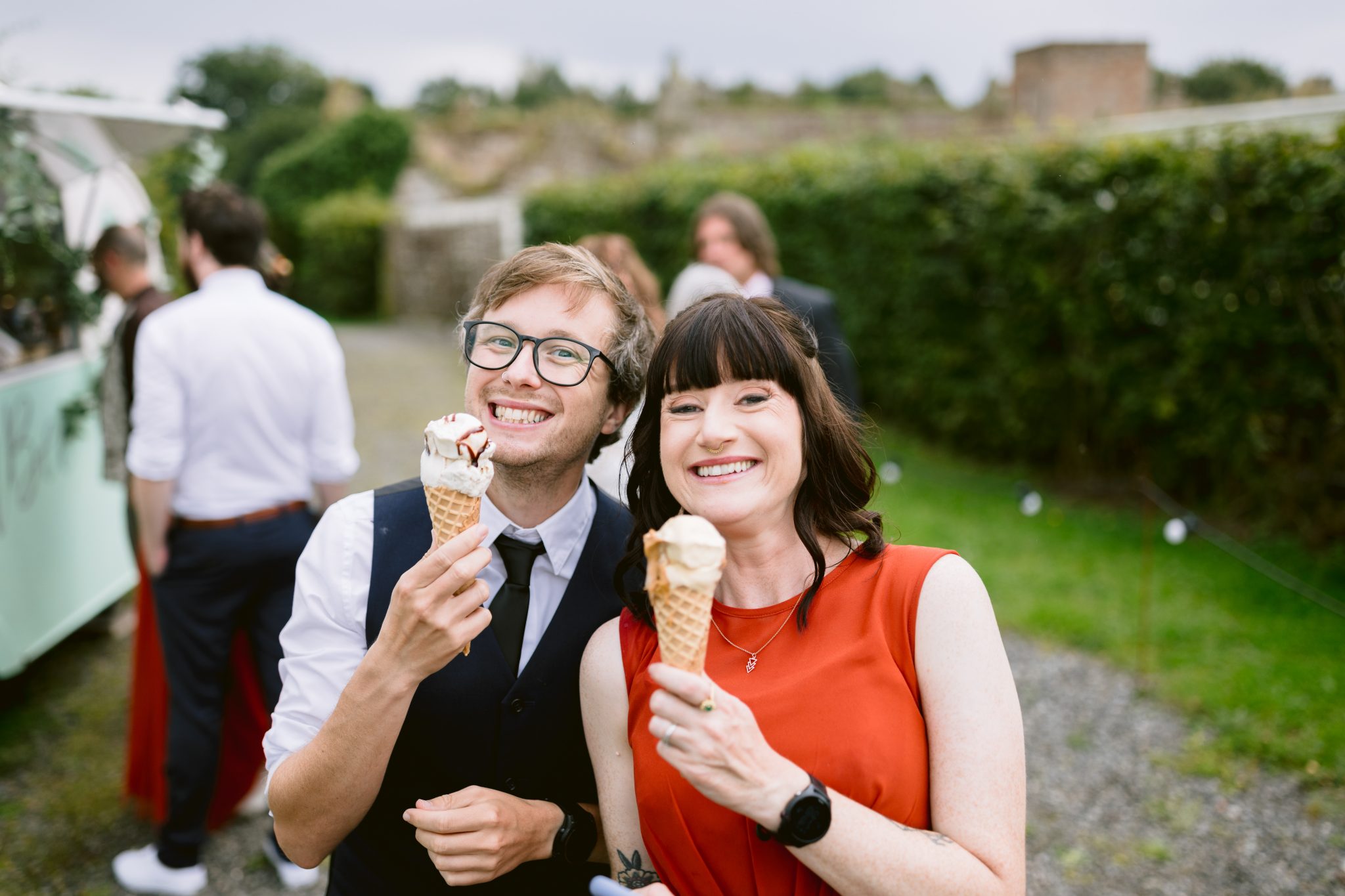 Two people smile as they enjoy ice cream cones amidst the lush greenery of Kirklinton Hall, with blurred figures in the background, capturing a perfect summer moment.