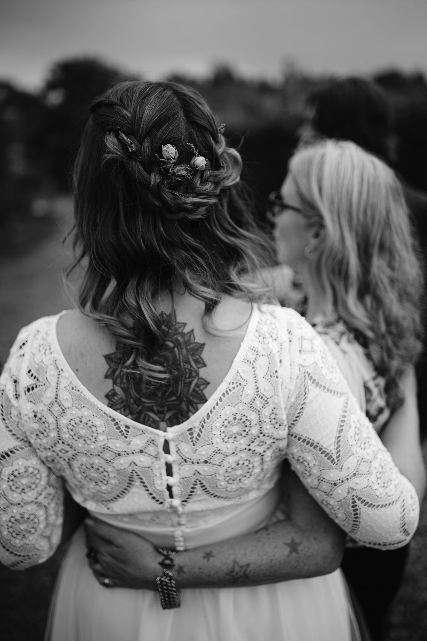 Two women with tattoos stand arm in arm amidst the lush gardens. One wears a lace dress, her hair intricately braided and adorned with flowers, as if ready for a Summer Marquee Wedding at Kirklinton Hall. The scene is set beautifully outdoors.