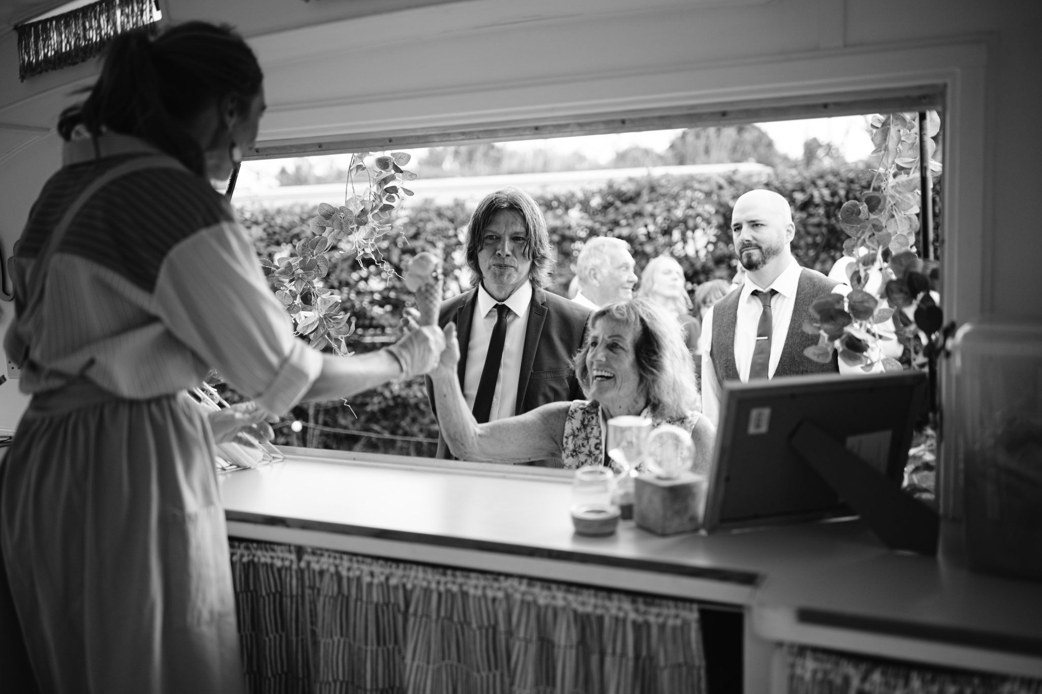 At a bustling food truck, parked just beyond the Gardens of Kirklinton Hall, a smiling woman is served by another inside. Behind her, several people, including two men in suits likely from a nearby Summer Marquee Wedding, wait patiently for their turn to savor the treats.