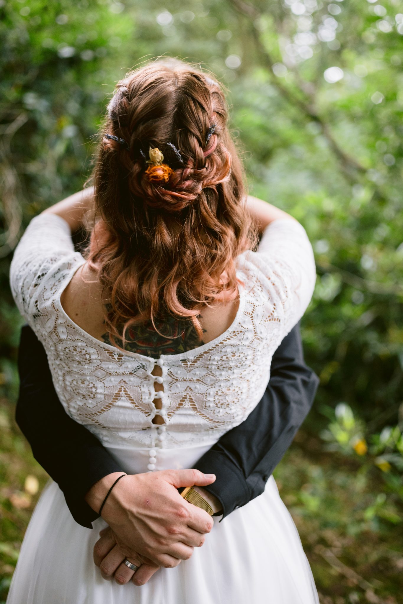 In the enchanting gardens of Kirklinton Hall, a couple embraces. The woman, in a white lace dress with braided hair adorned with flowers, faces away. The man, clad in a black suit, gently holds her waist against the backdrop of their summer marquee wedding.