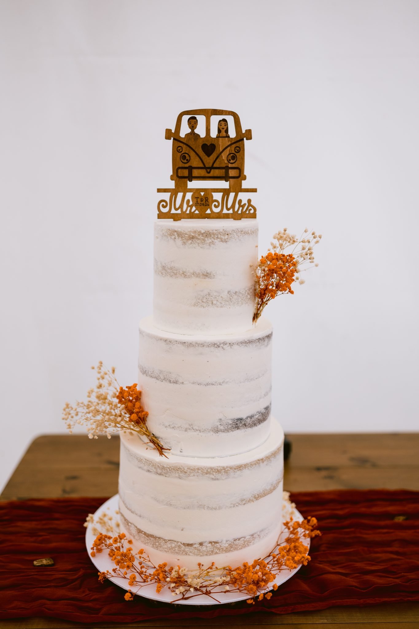 A three-tier white cake with minimal frosting and dried orange flowers, topped with a wooden "Mr and Mrs" sign featuring a car design, sits elegantly on a red cloth-covered table, perfectly capturing the charm of a summer marquee wedding at Kirklinton Hall.