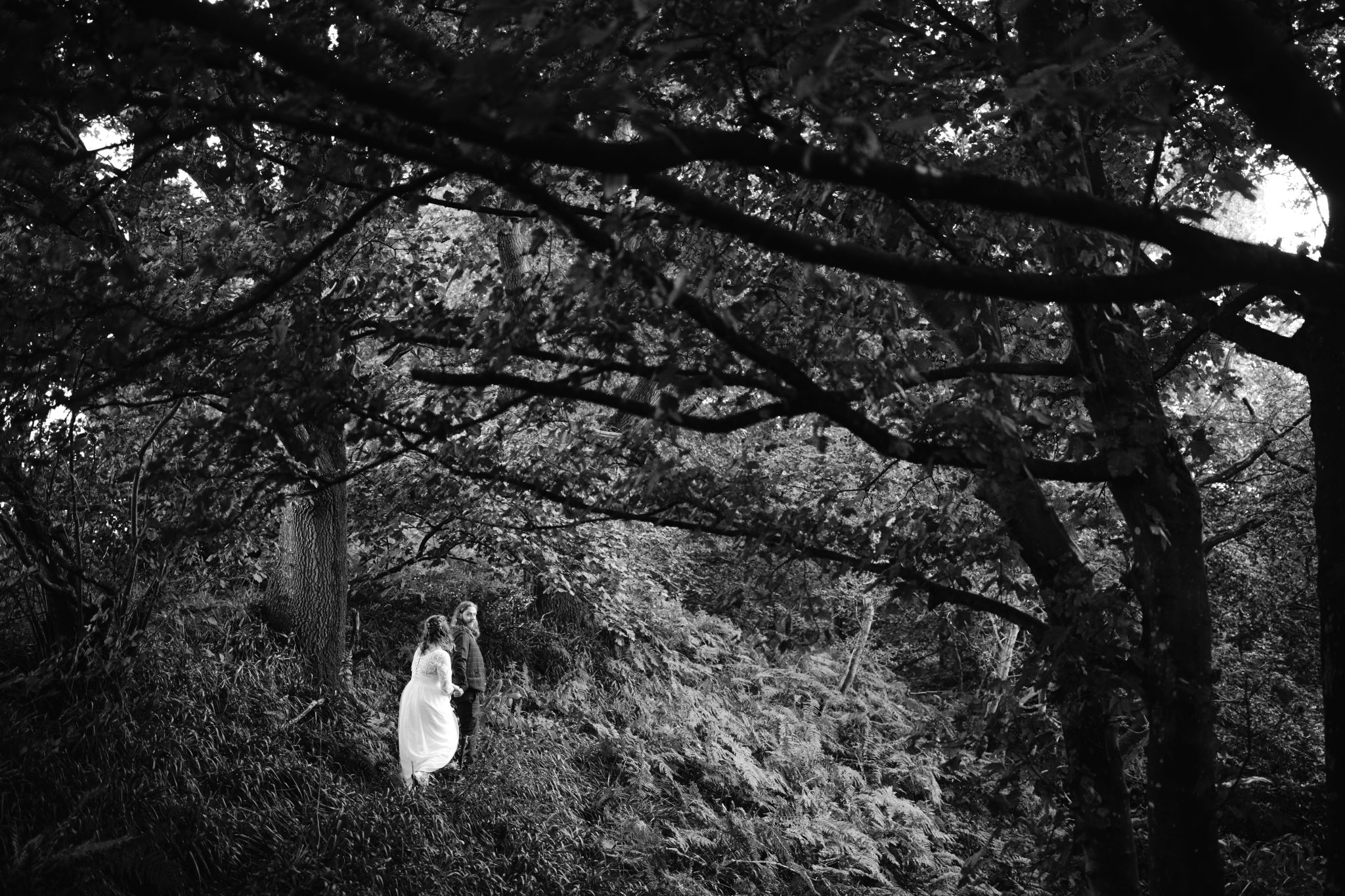 Two people stand on a forested hillside under large tree branches, one wearing a light-colored garment, reminiscent of an enchanting scene in the gardens of Kirklinton Hall. The black and white setting evokes the timeless elegance of a summer marquee wedding.