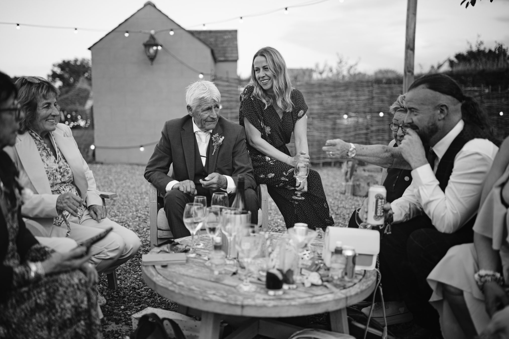 In the enchanting Gardens of Kirklinton Hall, a group of people in formal attire share conversation and laughter around a circular table with drinks. The Summer Marquee Wedding is beautifully illuminated by string lights in this picturesque outdoor setting.