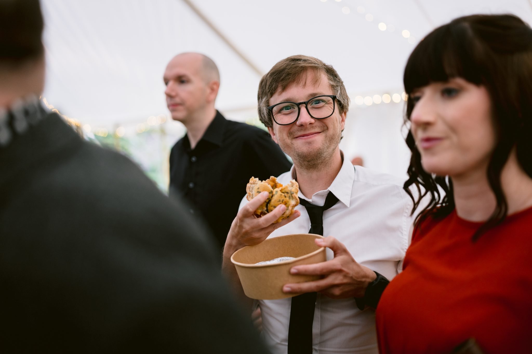A man in glasses, wearing a white shirt and tie, smiles while holding a bowl and sandwich. A woman in a red top and another man are visible against the backdrop of Kirklinton Hall's stunning Gardens.