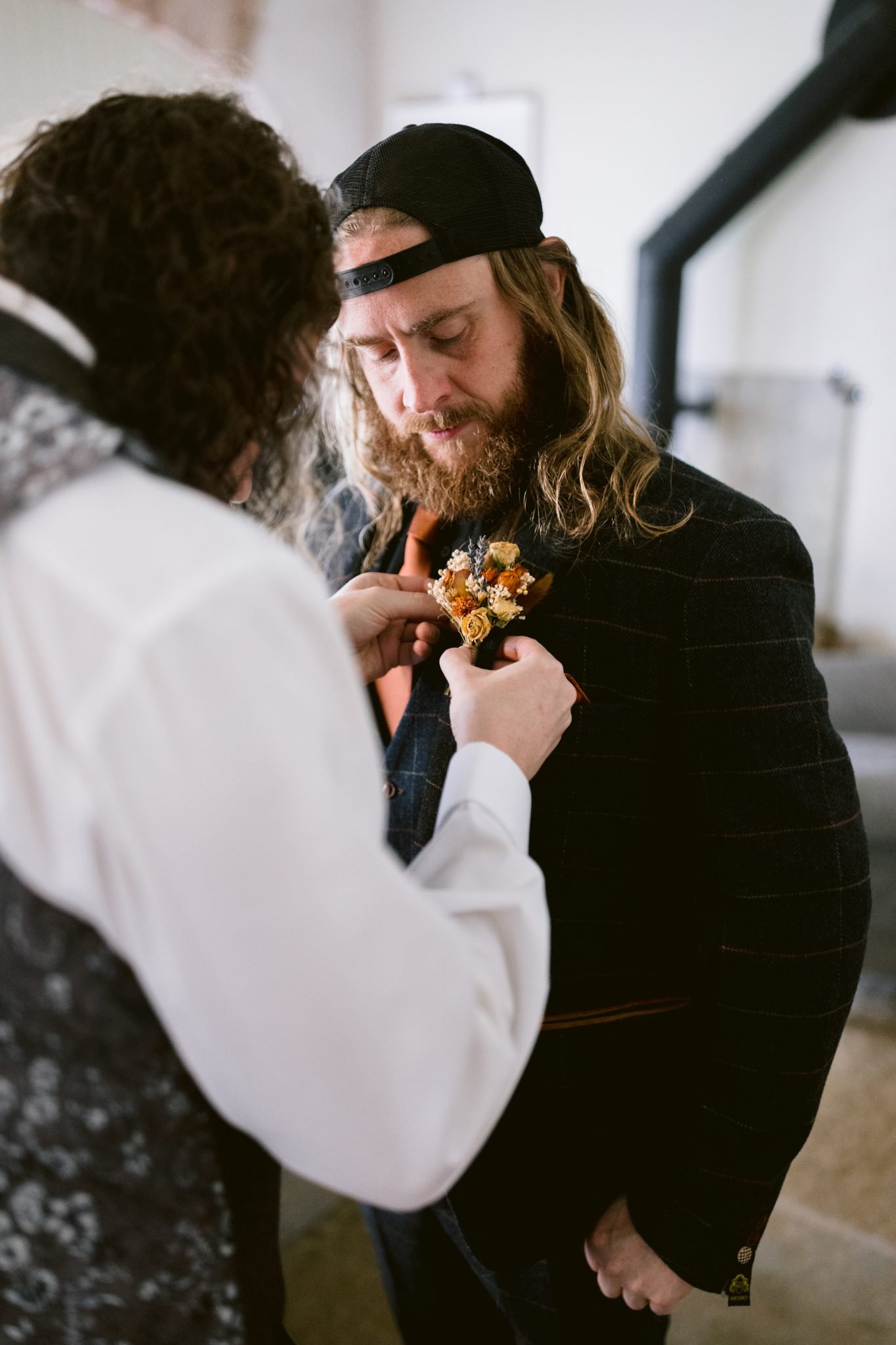 Indoors at Kirklinton Hall, a person in a suit is being assisted by another in adjusting a boutonniere on their jacket, preparing for a beautiful summer marquee wedding.