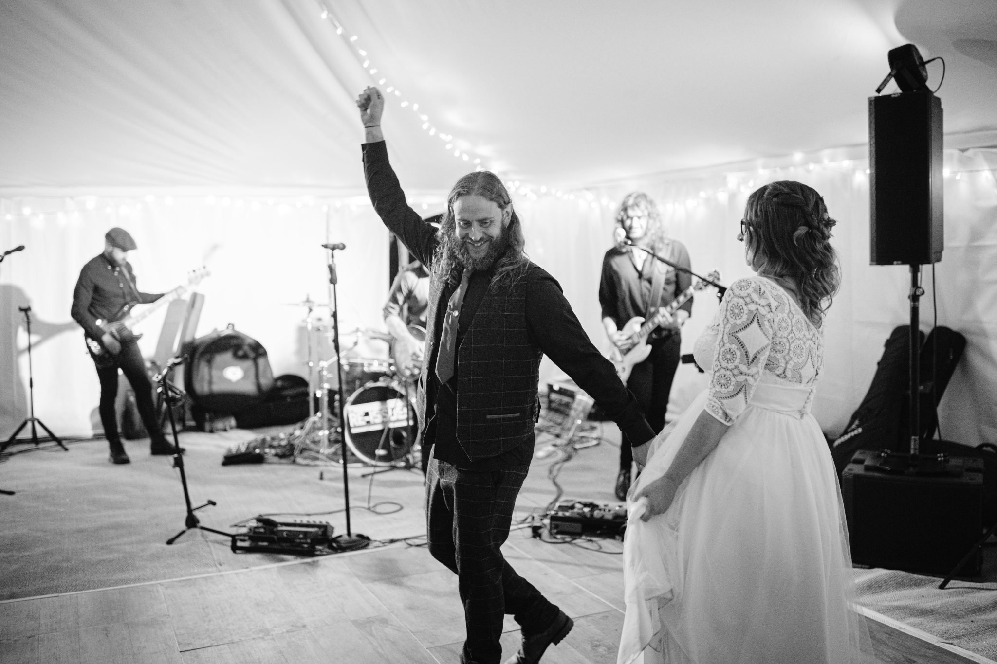 A man with long hair dances with a woman in a white dress at a summer marquee wedding. A band plays in the background under string lights, capturing the joy of the moment at Kirklinton Hall's gardens. Black and white photo.