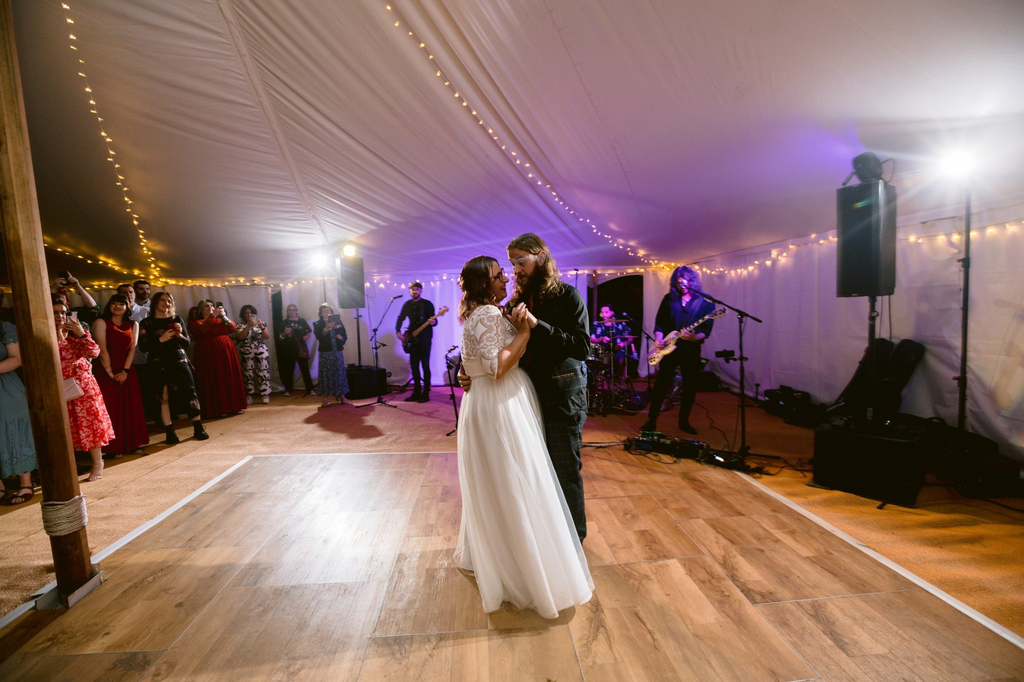 The bride and groom share a romantic first dance under twinkling string lights in a beautiful summer marquee wedding at Kirklinton Hall, surrounded by guests and the enchanting melodies of a live band performing in the gardens.