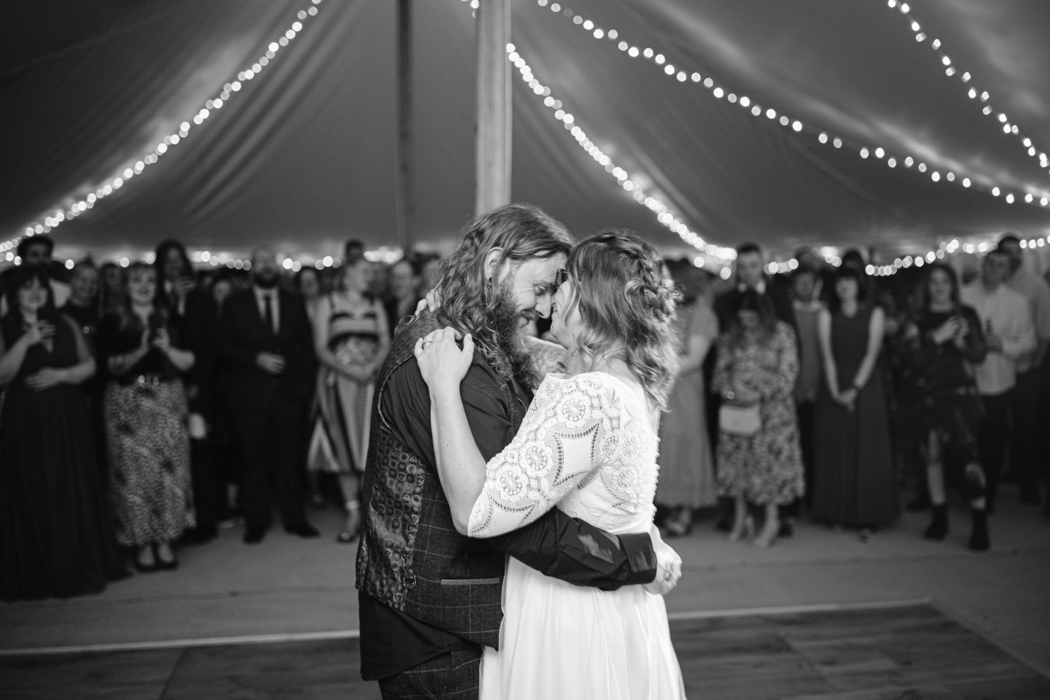 Amidst the enchanting setting of a Summer Marquee Wedding at Kirklinton Hall, a couple gracefully dances under string lights in a tent, with guests watching in admiration.