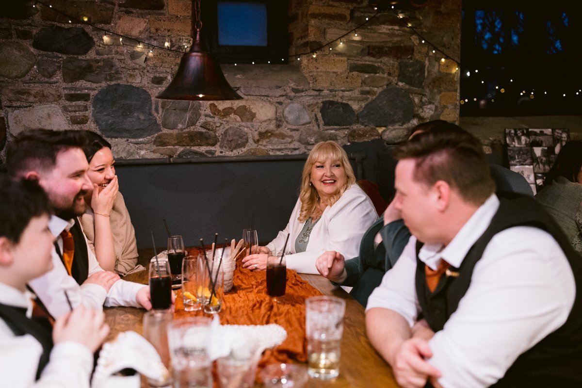 People sitting around a wooden table in a rustic setting, enjoying drinks at Low Hall The Lakes. A woman in white sits at the center, smiling. Strings of lights decorate the stone walls in the background, creating an enchanting atmosphere reminiscent of a small outdoor wedding.