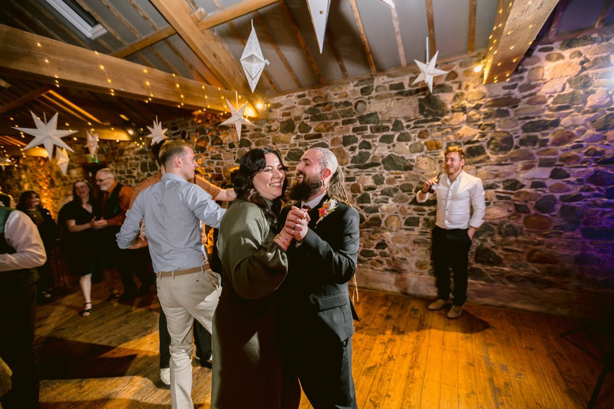 A couple dances in a rustic venue at Low Hall The Lakes with stone walls and wooden floors, surrounded by hanging star decorations and string lights. The intimate ambiance mirrors that of a small outdoor wedding, with other guests mingling in the background.