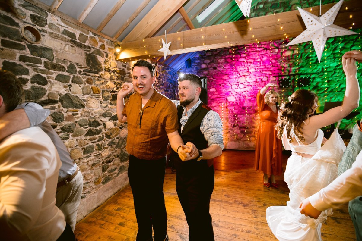 Two men in casual attire dance at an indoor event with a stone wall and colorful lights, reminiscent of vibrant nights at Low Hall The Lakes. Others join in the lively spirit, dancing blissfully in the background.
