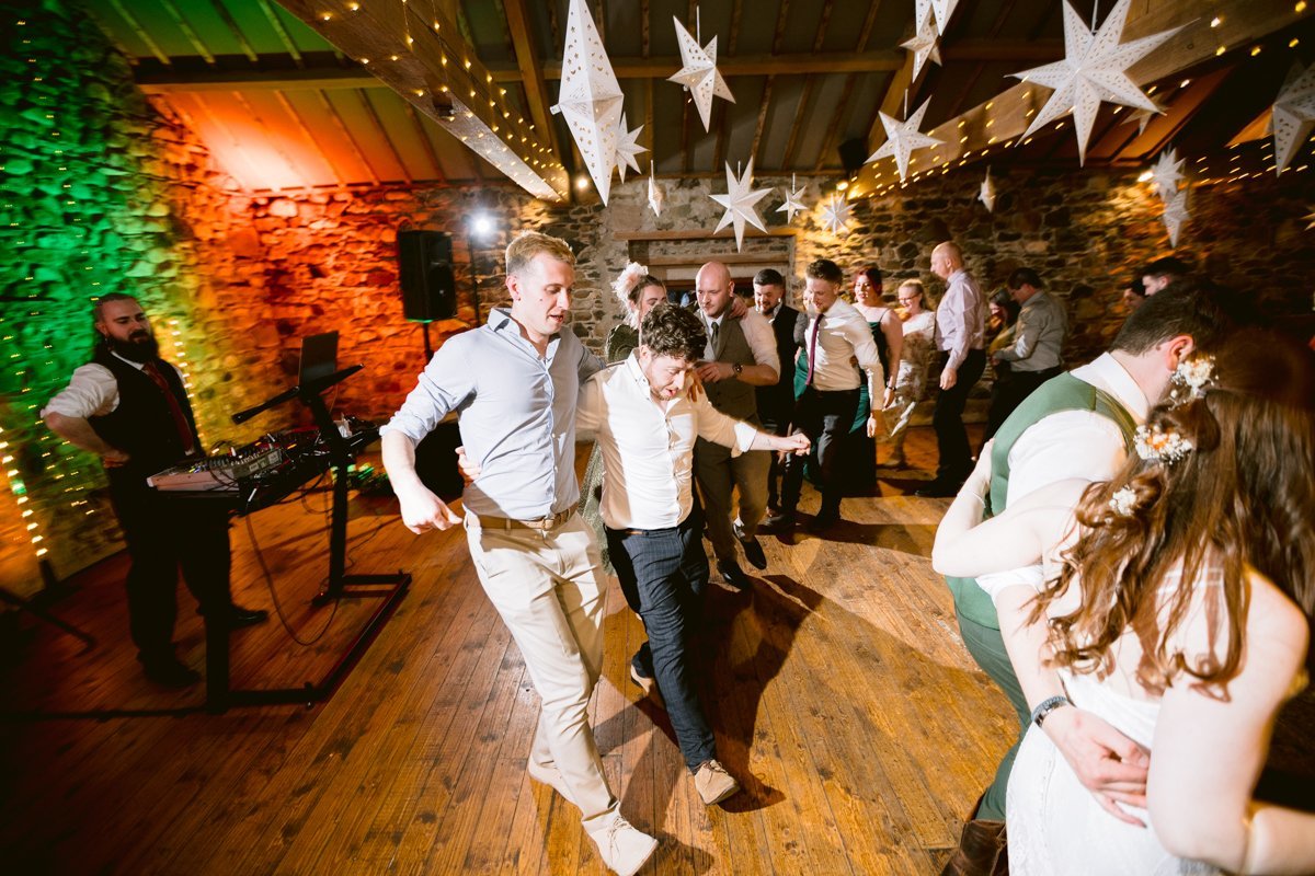 The rustic room feels alive as people dance energetically beneath star decorations, their feet tapping on the wooden floor. At Low Hall The Lakes, a DJ stations near speakers on the left, creating an unforgettable atmosphere reminiscent of a small outdoor wedding celebration.