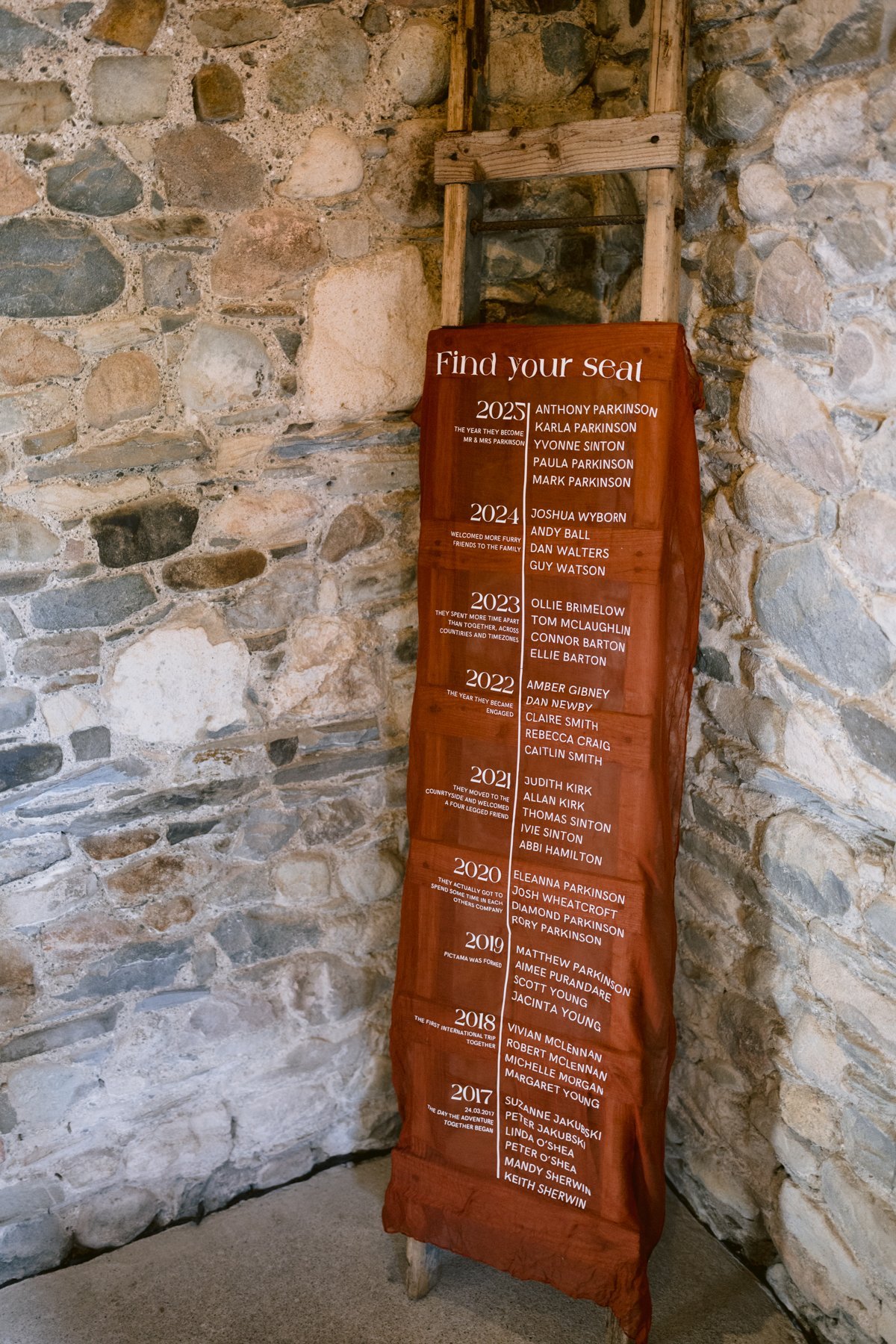 A wooden ladder draped with burgundy fabric lists names and years from 2017 to 2023, creating an elegant touch for a Low Hall The Lakes small outdoor wedding, all set against a rustic stone wall.