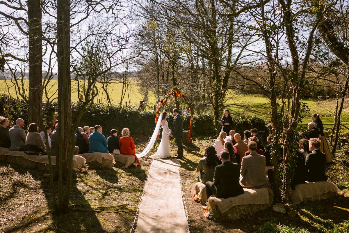 A small outdoor wedding unfolds in the serene beauty of The Lakes, with a couple standing under a floral arch. Guests sit on stone benches along a central pathway, surrounded by the tranquil charm of trees and fields.