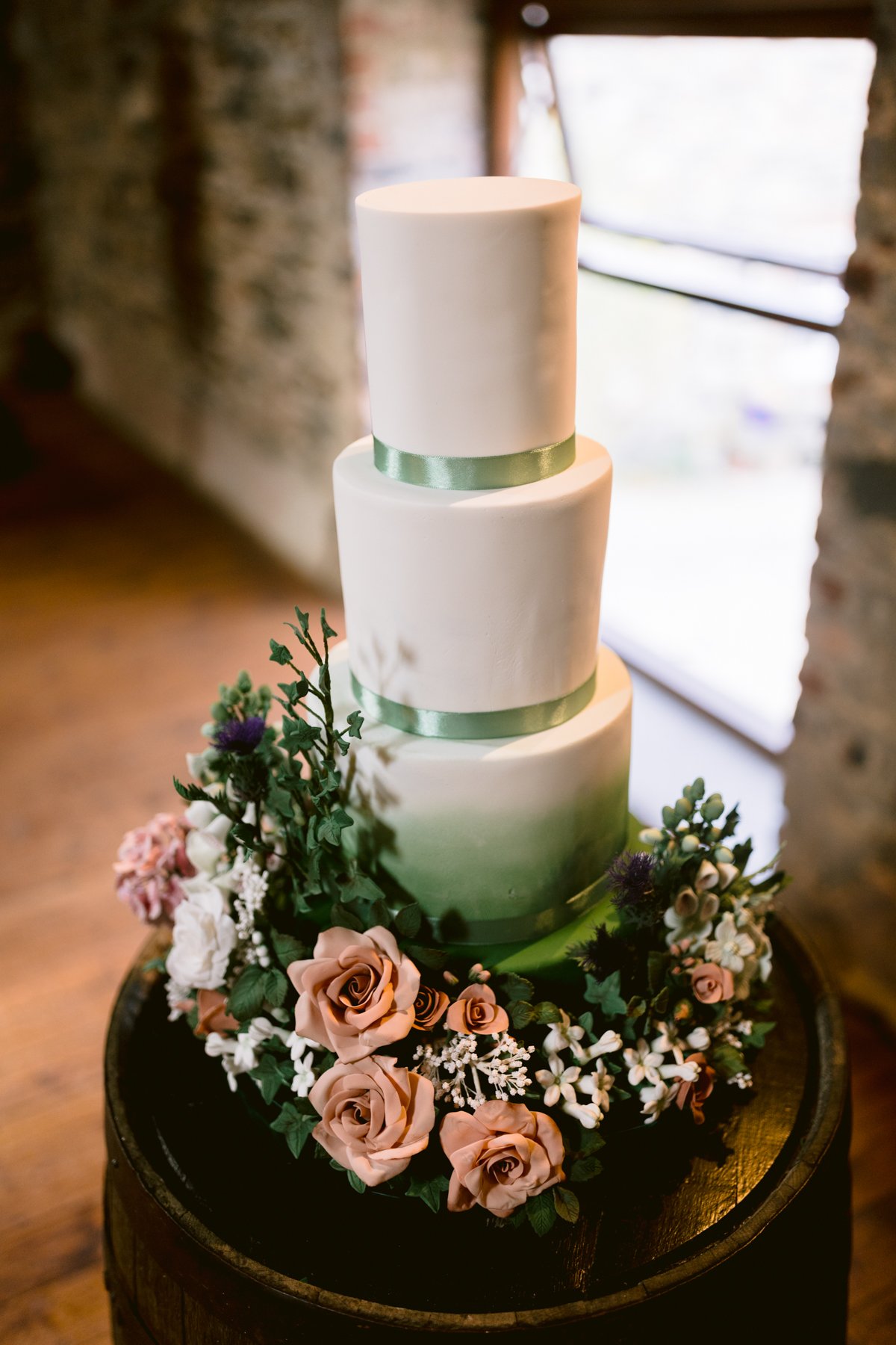 A three-tier white cake with green ribbon accents is beautifully adorned with pink and white flowers and lush greenery, set on a barrel in the rustic charm of The Lakes for an intimate small outdoor wedding.
