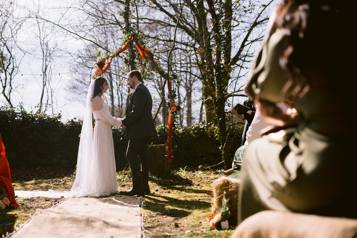 At a small outdoor wedding at Low Hall The Lakes, the bride and groom hold hands amidst the scenic beauty. Guests are seated nearby, all set against the backdrop of a stunning floral arch.