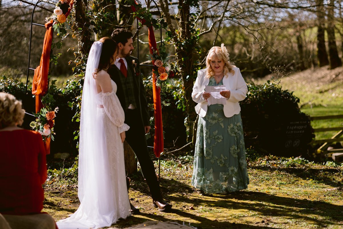 Under the floral arch at Low Hall The Lakes, a couple stands together, listening intently to a woman reading from a paper during their intimate outdoor wedding ceremony.