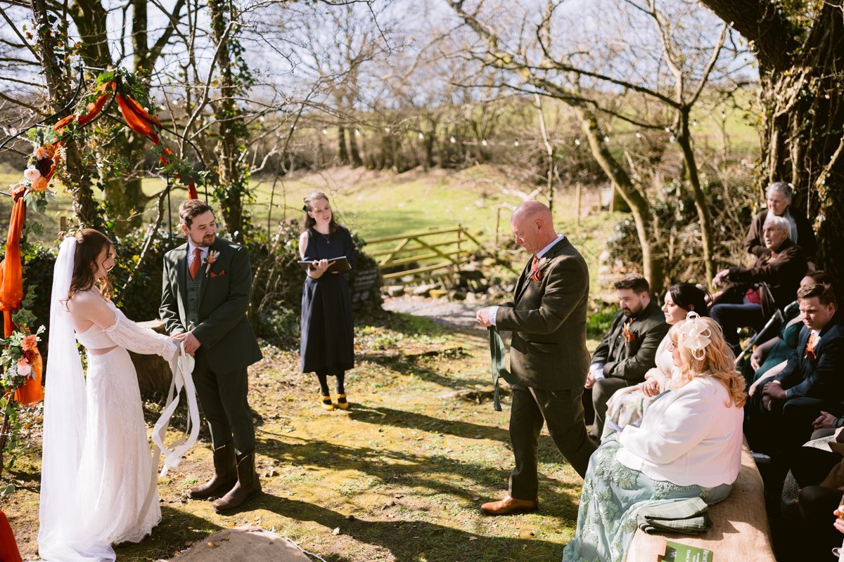 A couple, dressed in wedding attire, stands at their small outdoor wedding surrounded by rustic decorations. Gathered at Low Hall The Lakes, a person officiates the ceremony while others watch attentively amid the serene embrace of nature and towering trees.