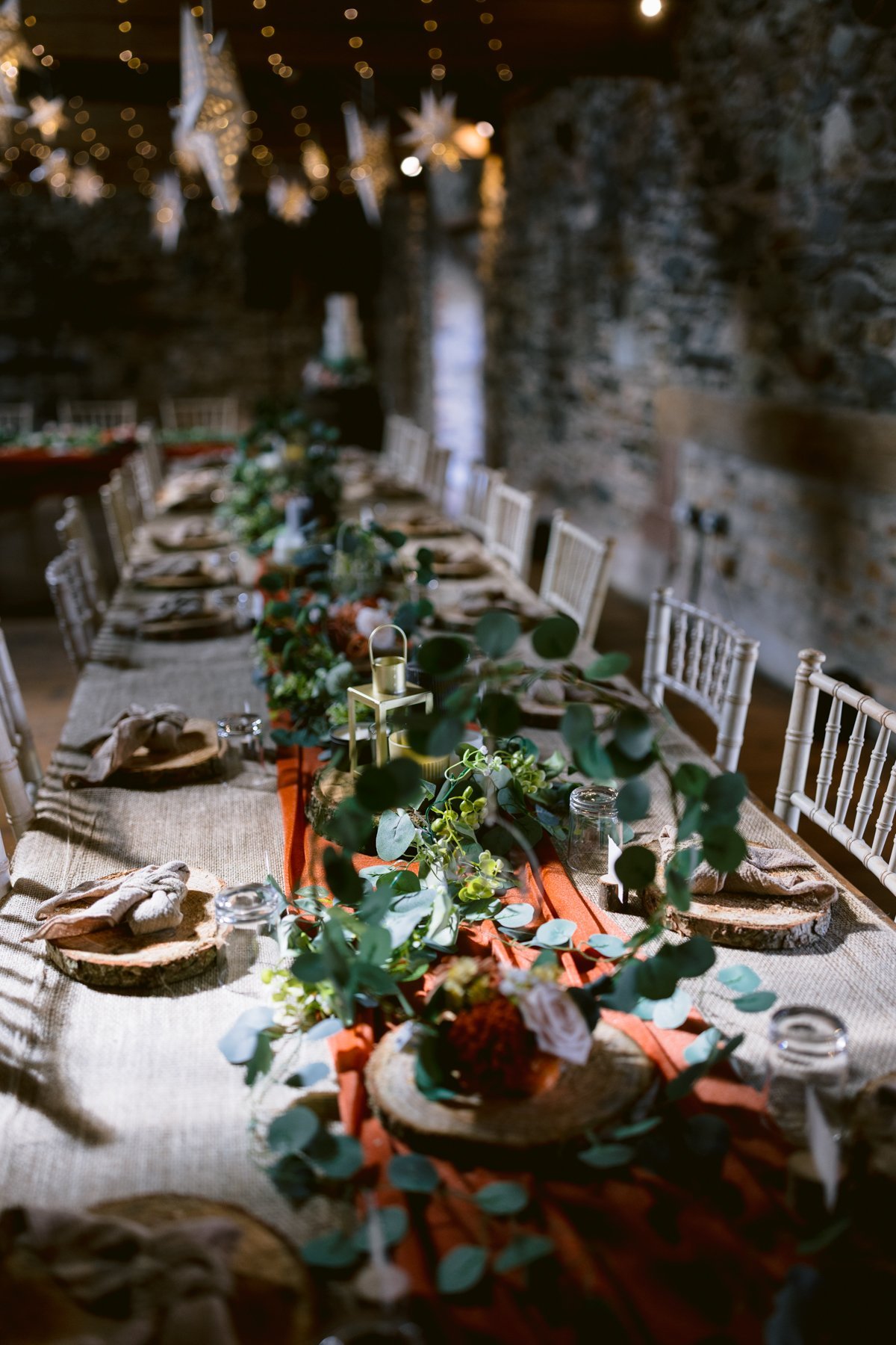 A rustic dining table is elegantly set for a meal, perfect for a small outdoor wedding at Low Hall The Lakes. With wooden chargers, green foliage, candles, and chairs, the stone wall and hanging lights create a cozy atmosphere.