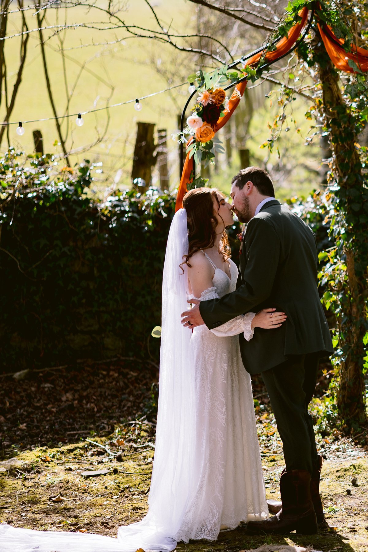 The bride and groom share a kiss outdoors at Low Hall, standing on a white carpet under a floral arch adorned with string lights. This idyllic setting perfectly captures the intimate charm of their small outdoor wedding by The Lakes.