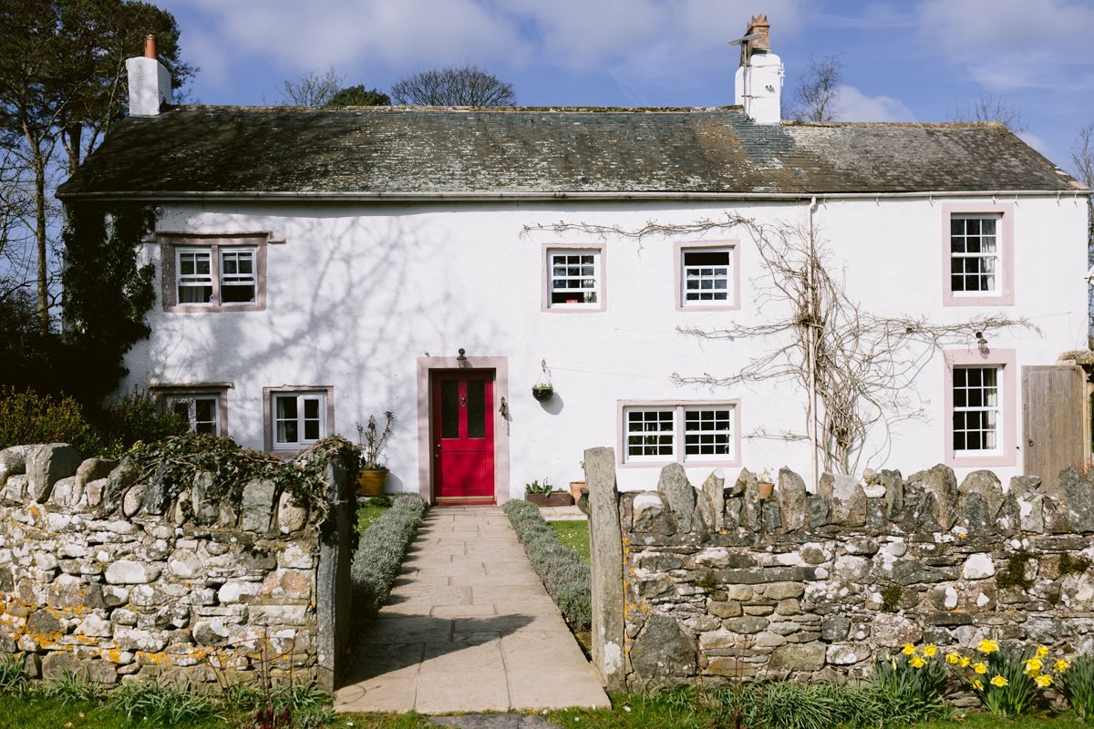 Nestled in nature, this old white cottage with a red door is perfect for a small outdoor wedding. Encircled by a stone wall and vibrant daffodils, its charming garden offers a picturesque setting against the backdrop of blue skies and whispering trees at Low Hall The Lakes.