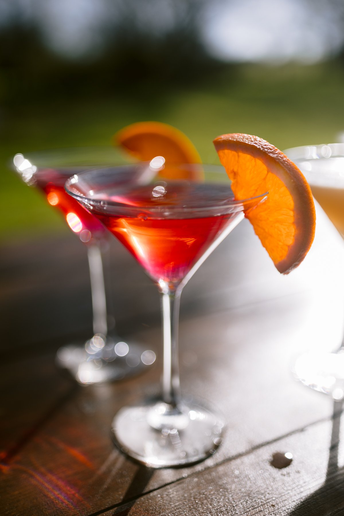 On a wooden table bathed in sunlight, three cocktail glasses with red drinks and orange slices capture the essence of a perfect outdoor wedding at Low Hall The Lakes.
