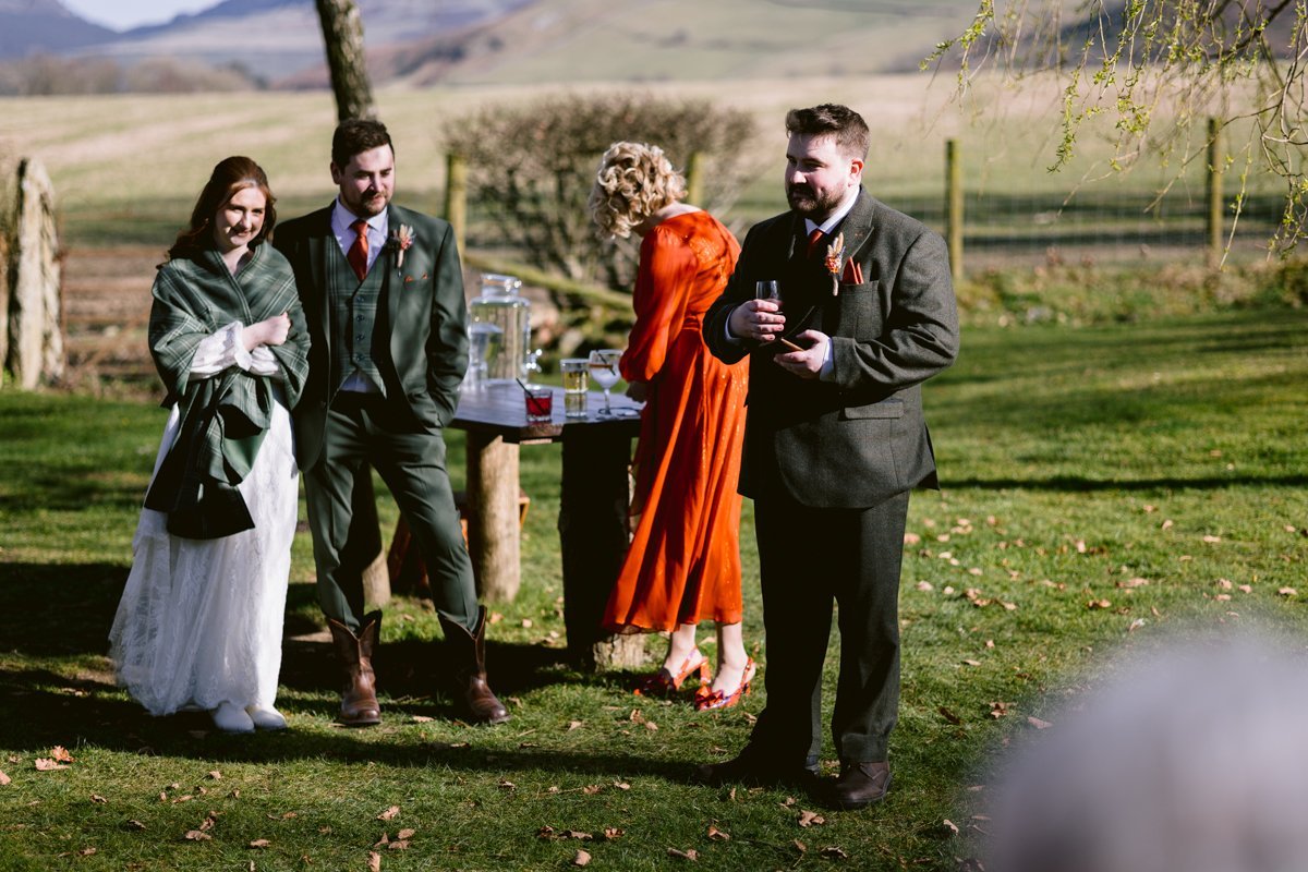 A group gathers on the grass in the countryside for an intimate outdoor wedding at Low Hall The Lakes. One person wears a white dress and green shawl, another dons a suit with boots. A person in red pours a drink, while a man holds a cup.