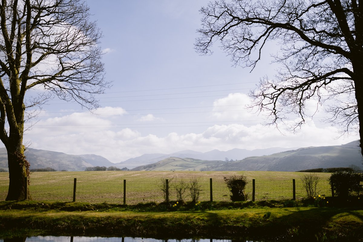 A serene landscape featuring a grassy field bordered by trees, a fence, and distant mountains under a partly cloudy sky provides the perfect setting for a small outdoor wedding at Low Hall The Lakes.