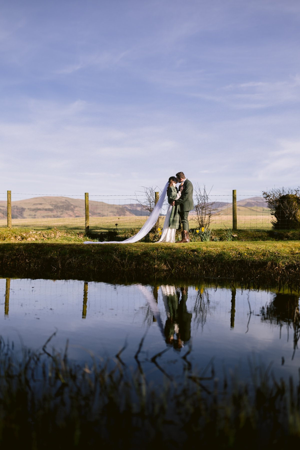 A couple in wedding attire embraces by a serene pond with reflections, celebrating their small outdoor wedding. The idyllic scene at Low Hall The Lakes features a fence and rolling hills under a clear blue sky.