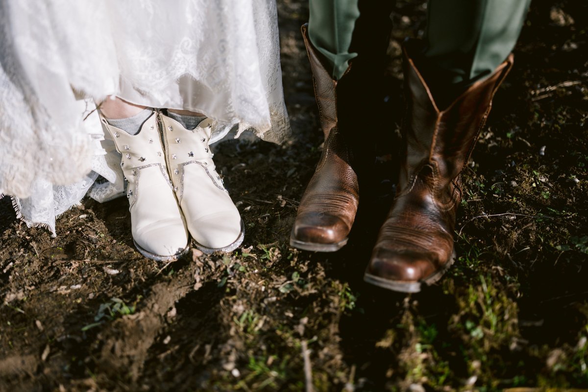 White bridal boots and brown cowboy boots standing on the dirt ground, perfect for a small outdoor wedding at Low Hall, The Lakes.