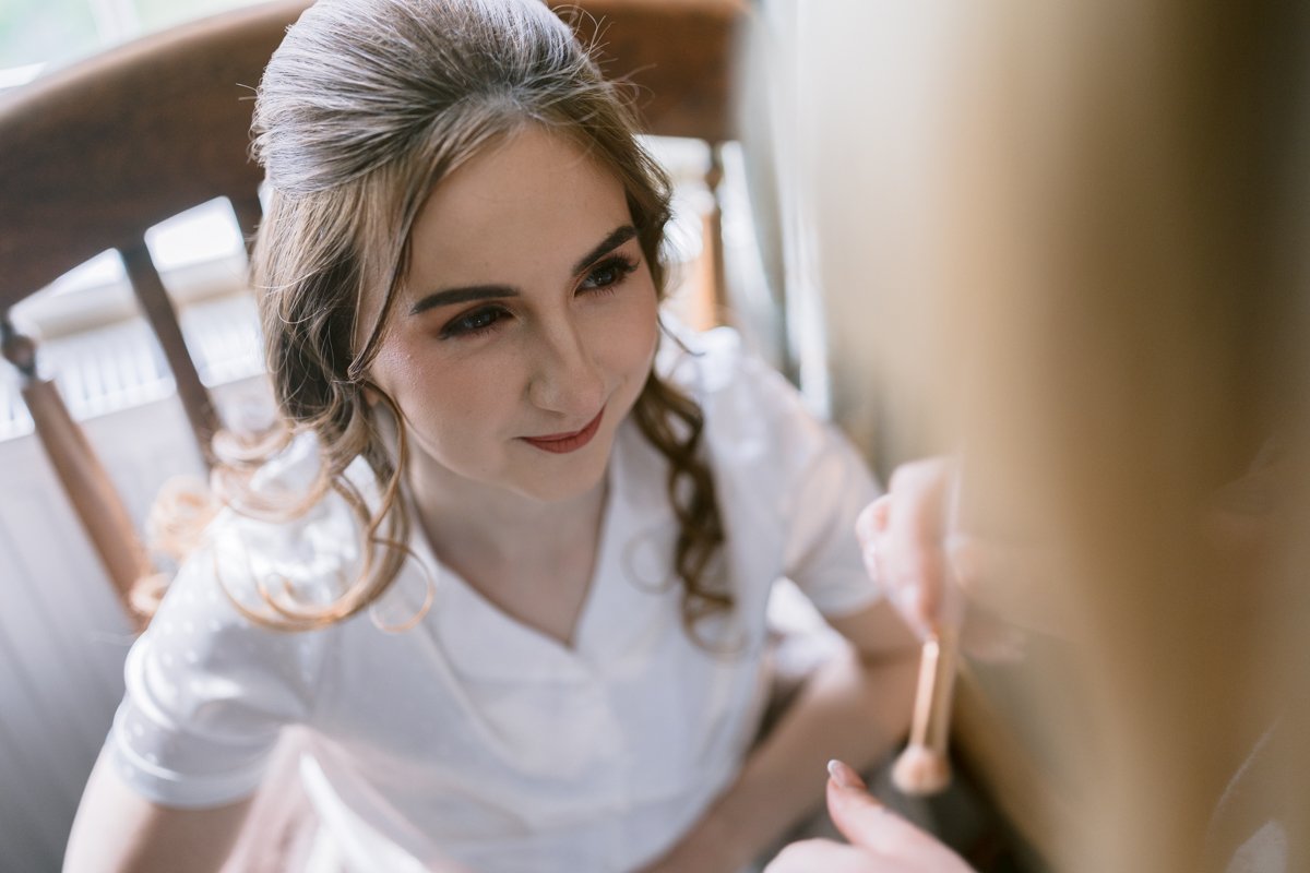 At Low Hall The Lakes, a woman with long hair sits in a chair wearing a white top, preparing for her small outdoor wedding as makeup is carefully applied to her face.