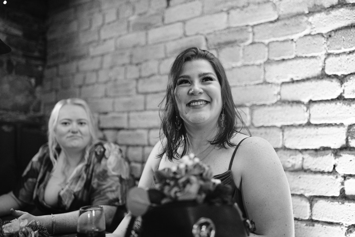 Two women sit in a restaurant with a brick wall backdrop, one smiling and looking up, the other with a neutral expression. A flower arrangement graces the foreground. The scene exudes an intimate charm reminiscent of a small outdoor wedding at Low Hall The Lakes. Black and white photo.