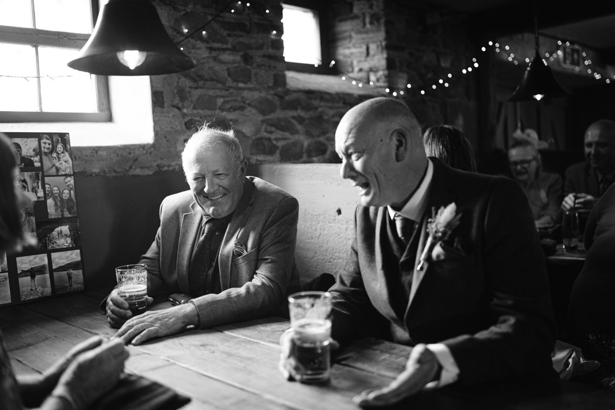 In the cozy ambiance of Low Hall The Lakes, two men in suits sit at a wooden table, surrounded by strings of lights. They hold drinks and smile, enjoying the intimate atmosphere of this small outdoor wedding.