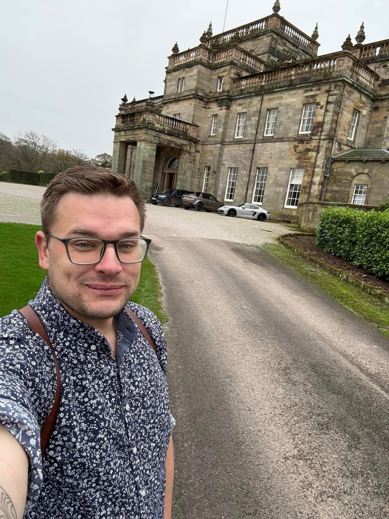 A person with glasses takes a selfie in front of a large historic building with cars parked nearby on a cloudy day.