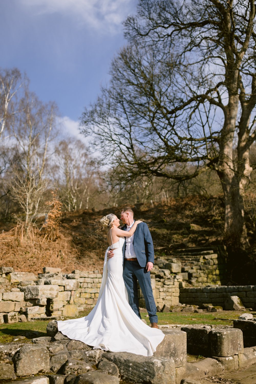 A bride and groom share a kiss outdoors among ancient stone ruins, with bare trees and a clear sky in the background.