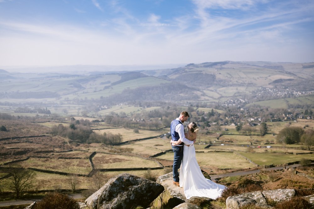 A bride and groom stand on a rock, embracing, with a scenic countryside landscape in the background under a blue sky.