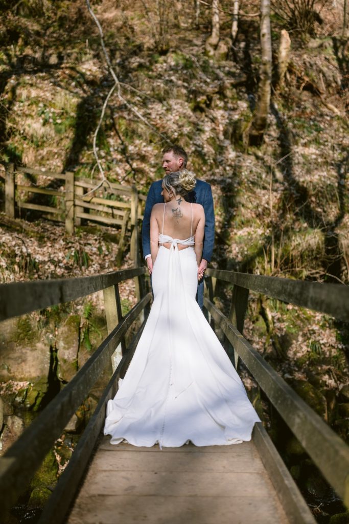 Bride and groom stand on a narrow wooden bridge surrounded by trees, with the groom facing the bride, who wears a white dress with an open back.
