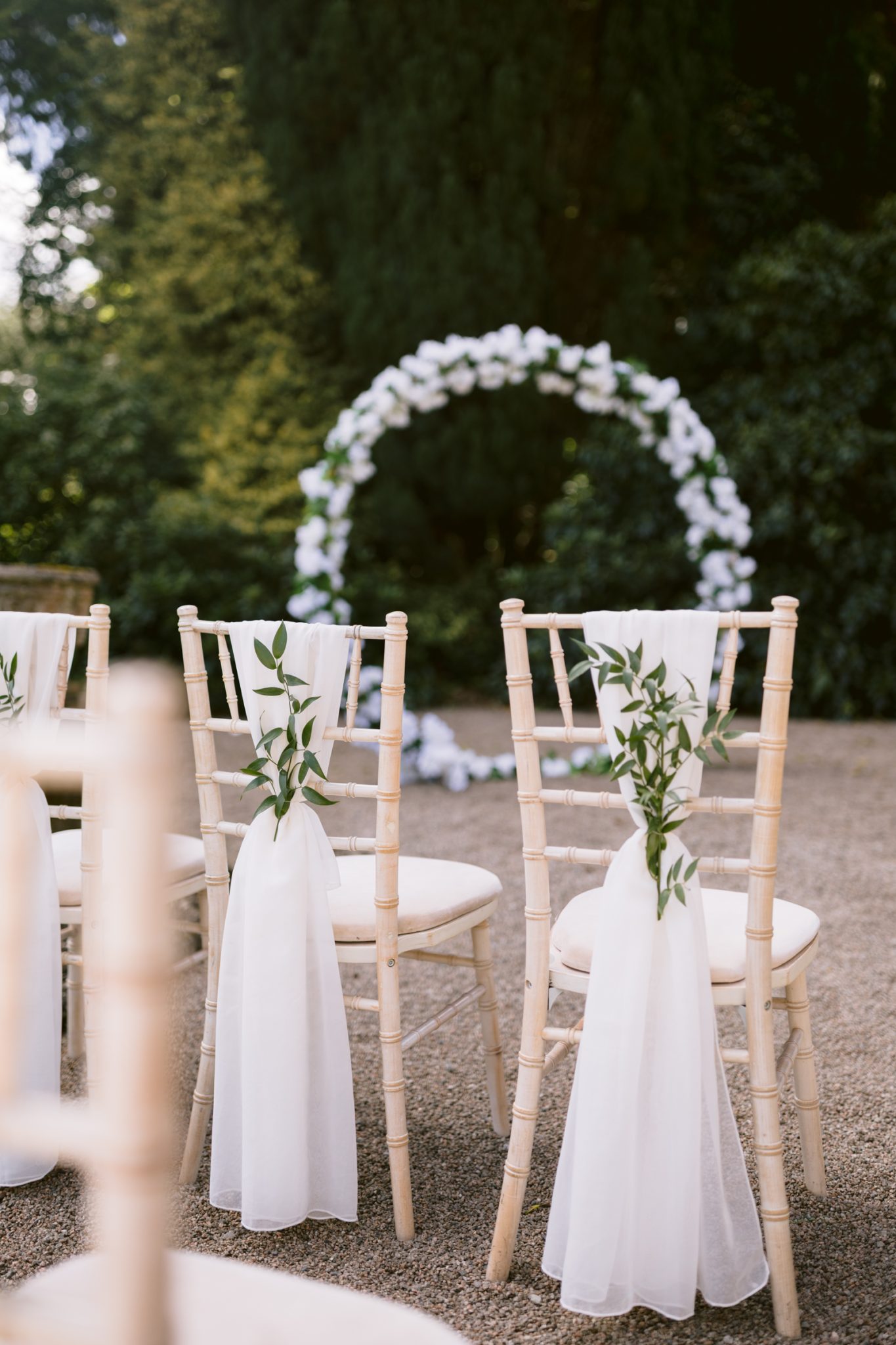 Rows of light wooden chairs with white fabric and greenery decorations face a white floral arch outdoors, set up for a wedding ceremony.