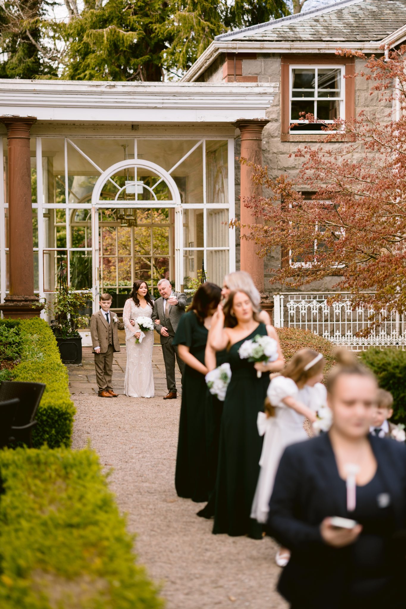 A bride and her party walk down a garden path toward a historic building, with bridesmaids and children leading the procession.