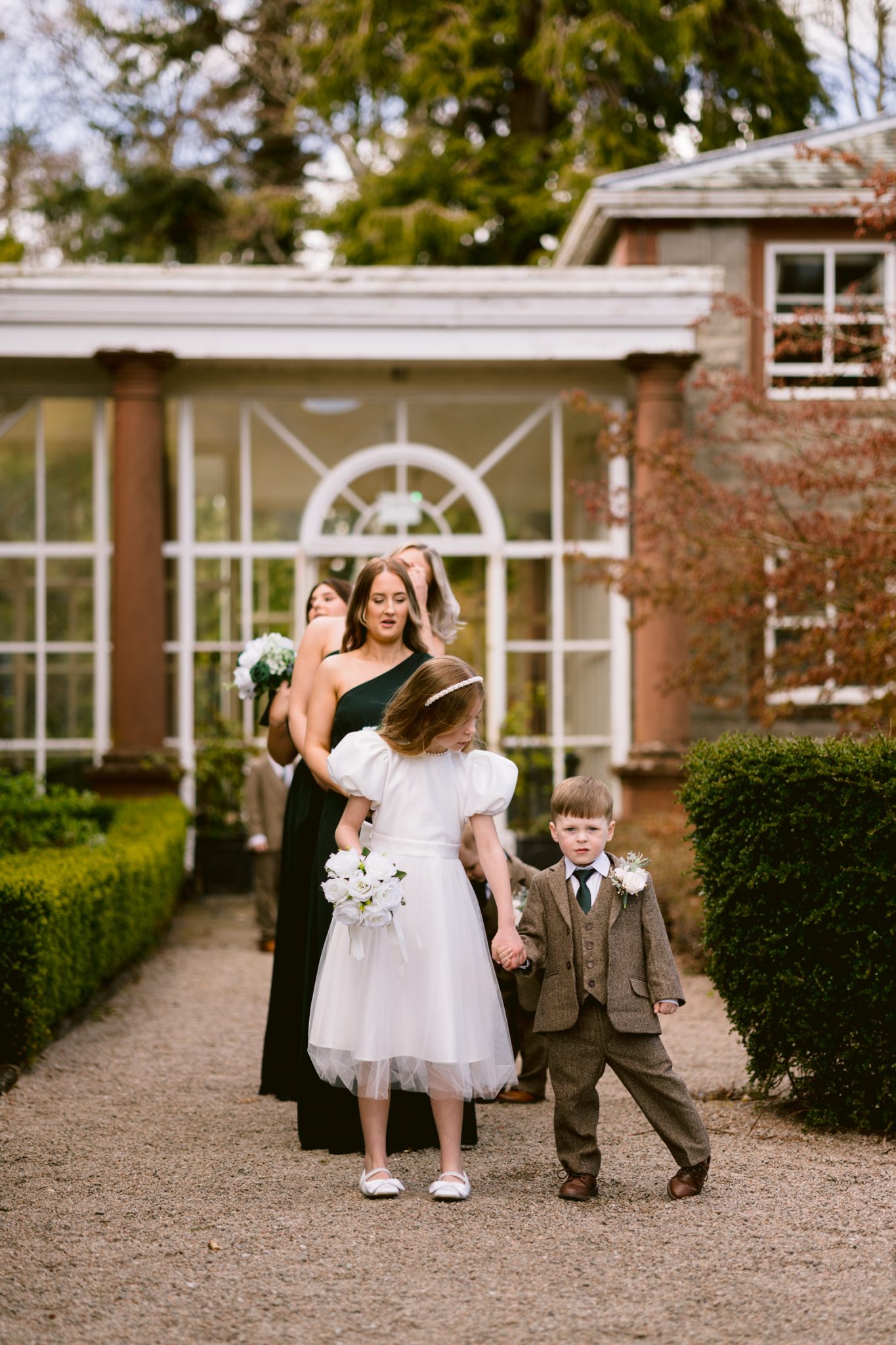 A girl in a white dress and a boy in a suit hold hands while walking outside, followed by women in dark green dresses, in a garden setting.